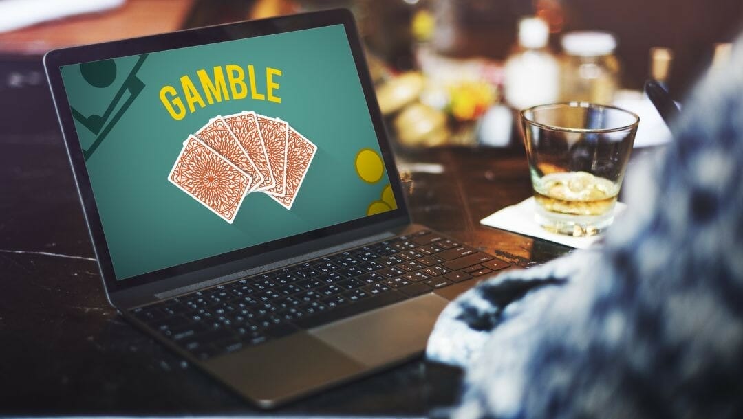 Body image, man on a laptop that displays a gambling concept with the word “GAMBLE” and five cards face down fanned out, drink beside the laptop 