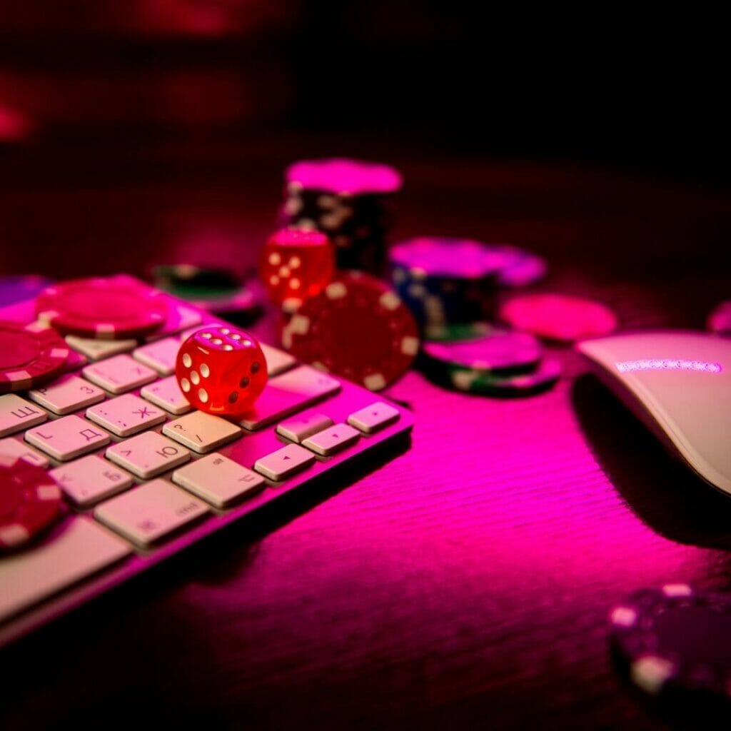 Poker chips and dice scattered around a keyboard and mouse with hot pink ambient lighting.