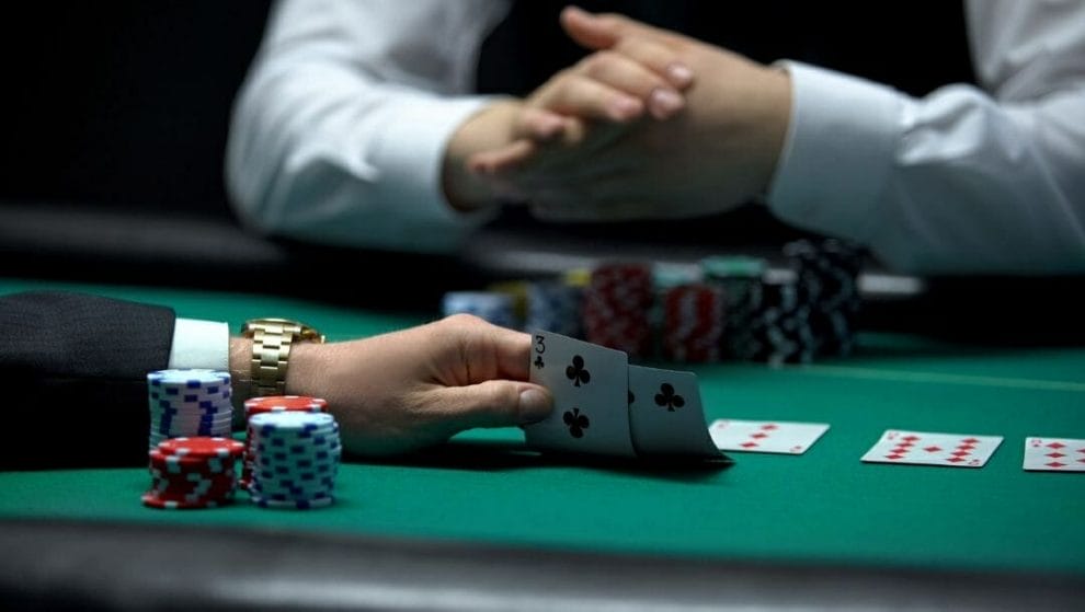 Body image, two men sitting opposite each other at a poker table, close up of their hands, the man closest to the camera is wearing a gold watch and checking his hole cards, the other man clasps his hands together, stacks of poker chips in front of each of them and playing cards lay face up on the table