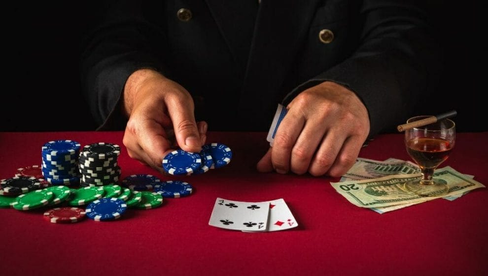 Body image, a man sits at a red poker table holding two poker chips out, stacks of poker chips to his right and a drink and money to his left, pocket fours lay face up on the table in front of him 
