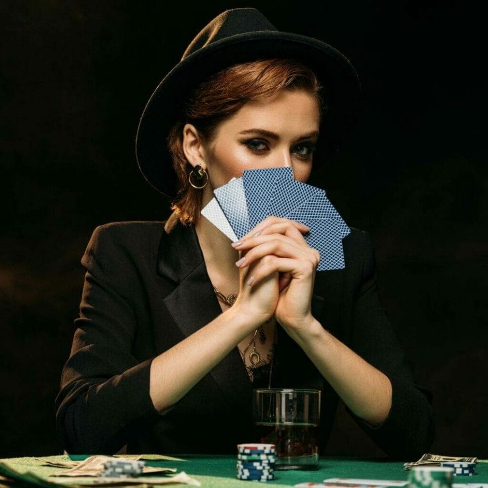 Header image, woman wearing a hat sitting at a poker table with a drink, poker chips and money in front of her, she holds up six playing cards in front of her face