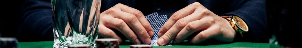 Hero, a man’s hands checking his hole cards at a poker table with a drink in front of him, he is wearing a gold watch on his left hand