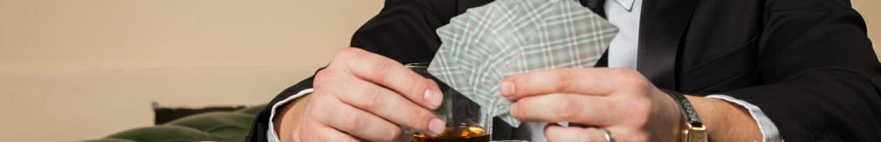 Hero image, close up of a man wearing a suit holding playing cards in his left hand and a drink in his right hand