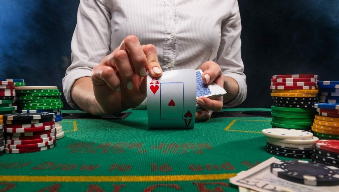 Croupier flipping up an ace of hearts towards the screen on a green blackjack table with stacks of poker chips surrounding the sides