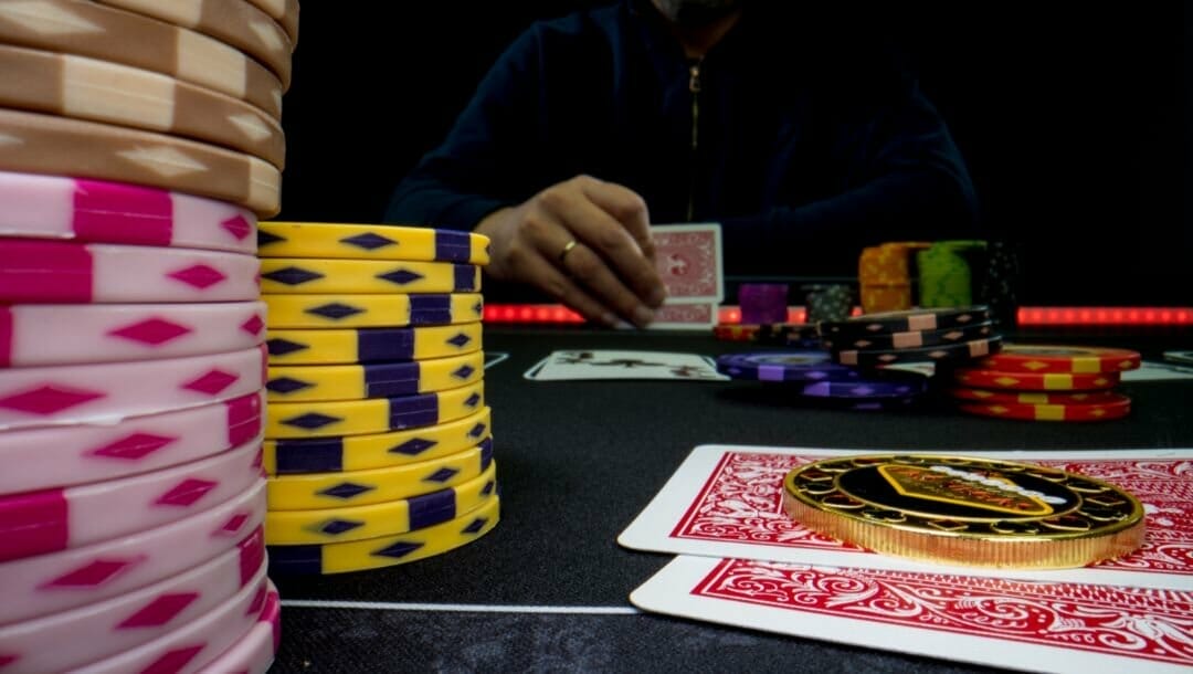 A person wearing a gold ring sitting at a poker table looking at their hole cards. The table is full of stacked poker chips and playing cards.