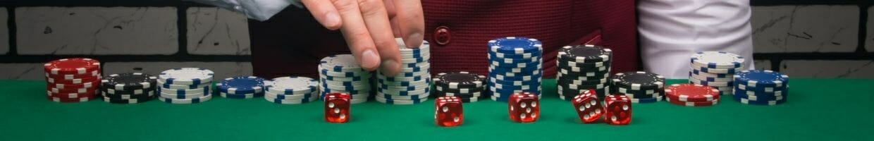 Stacks of poker chips on a green surface with five red dice in front of them. A person’s hand adjusts a stack of blue and white poker chips.