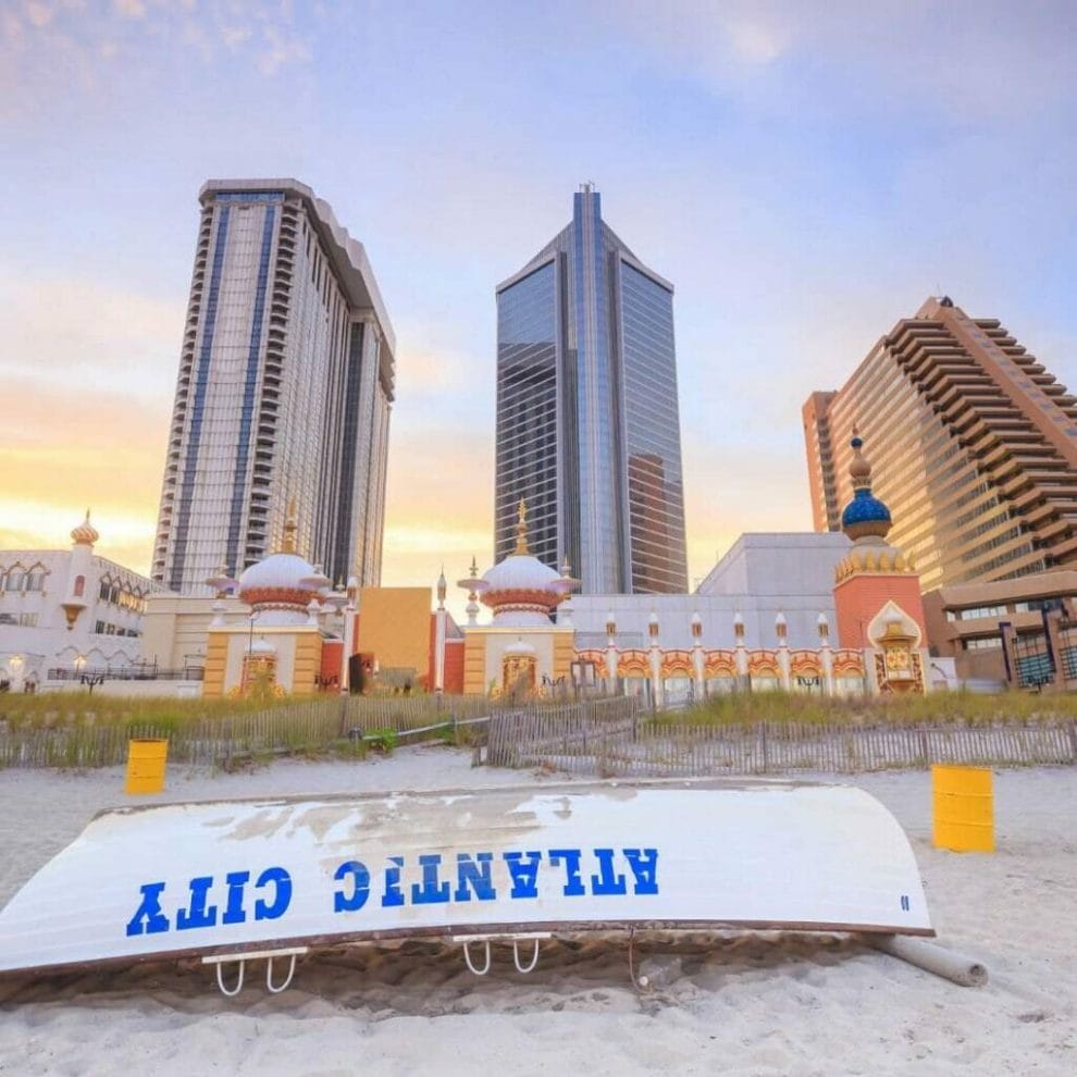 Header Image - Front shot of atlantic city casino resorts from the beach with an upside down rowboat on the sand that says “ATLANTIC CITY” on it, casino resort buildings behind
