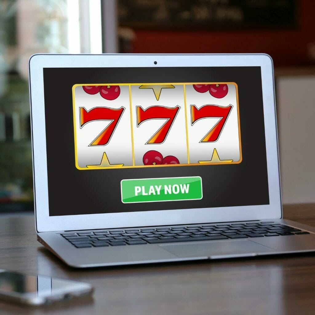 Header image, laptop open on a desk displaying online slot machine with a phone laying on the table next to the laptop