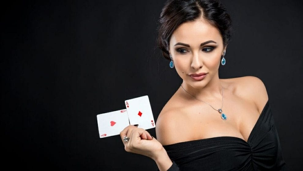 A woman holds up a pair of aces playing cards. She is wearing a black dress and jewelry set with blue stones.
