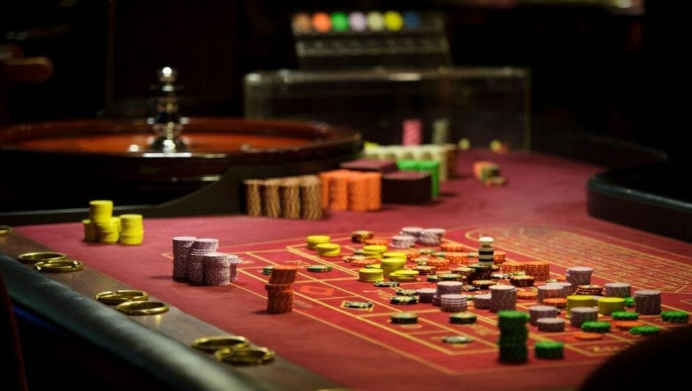 a red felt roulette table stacked with poker chips and a roulette wheel in the background inside a casino 