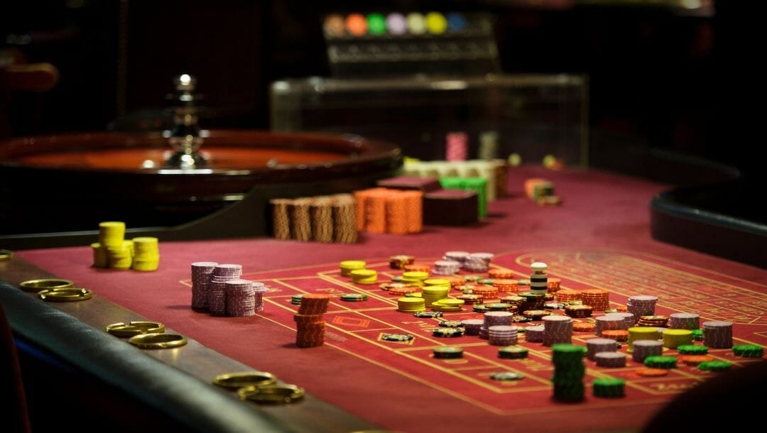 a red felt roulette table stacked with poker chips and a roulette wheel in the background inside a casino 
