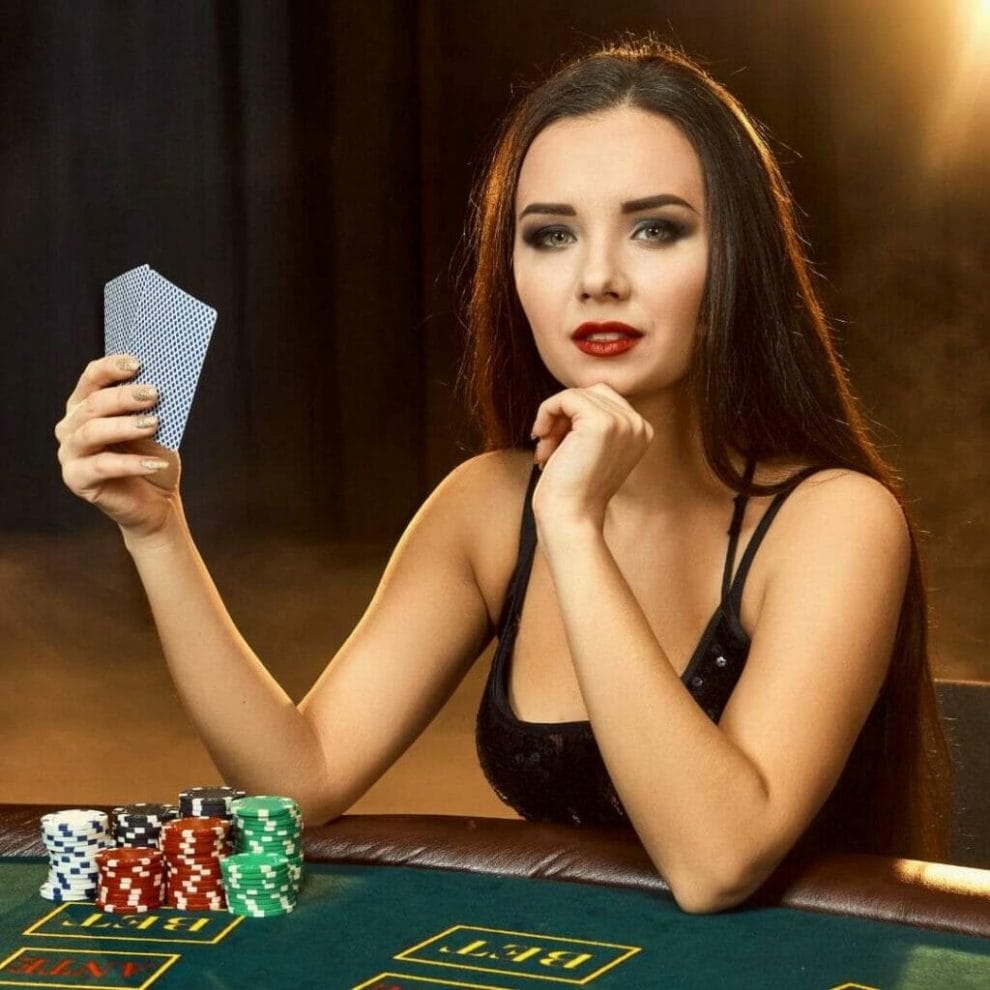 a brunette woman with long hair wearing a black sequin dress plays poker at a green felt poker table with playing cards in her hand and stacks of poker chips on the table in front of her