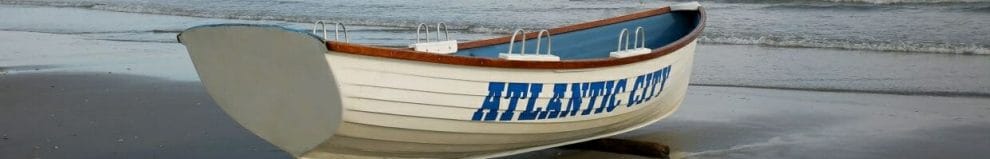 a white rowboat with the words “ATLANTIC CITY” painted on in blue on the shore with ocean waves in the background