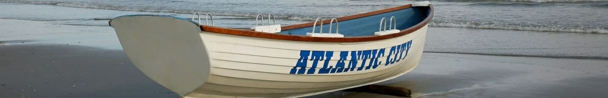 a white rowboat with the words “ATLANTIC CITY” painted on in blue on the shore with ocean waves in the background