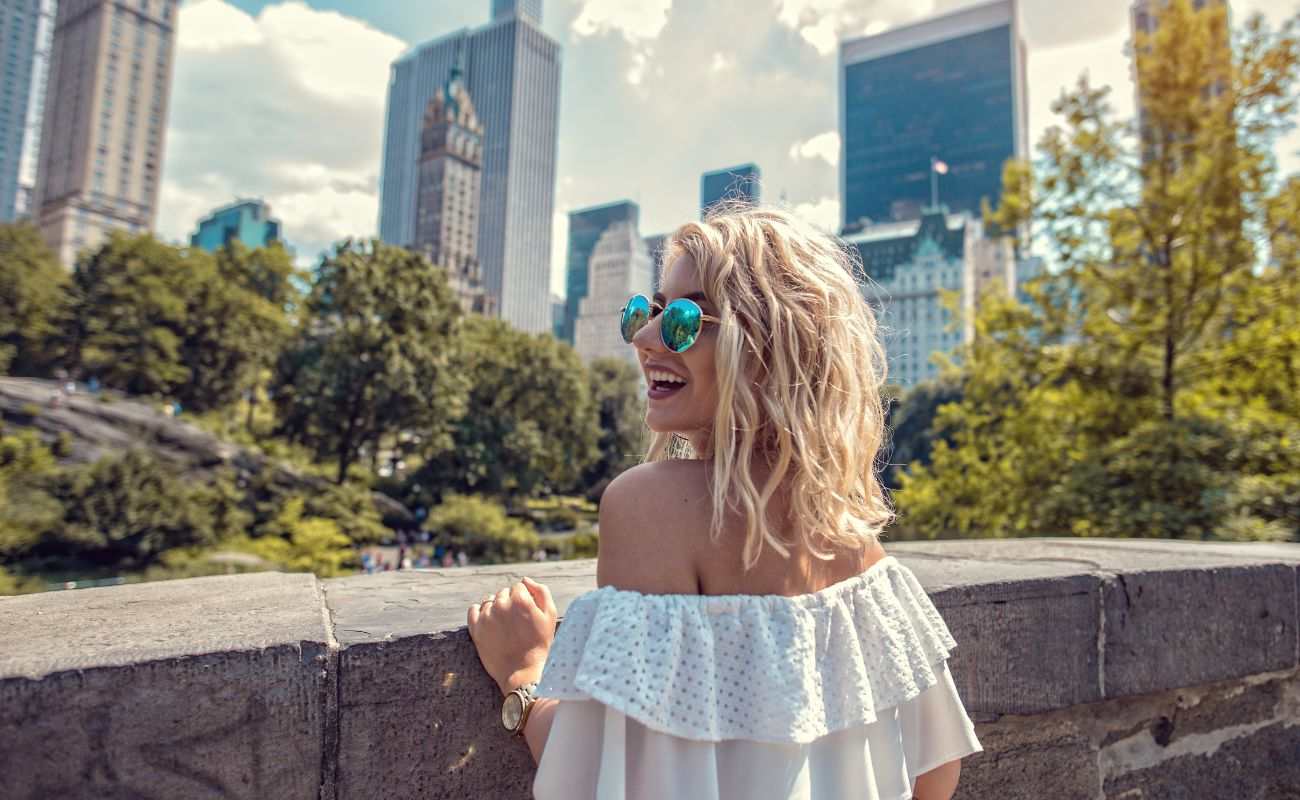 a woman poses in Central Park with the Manhattan skyline behind her 