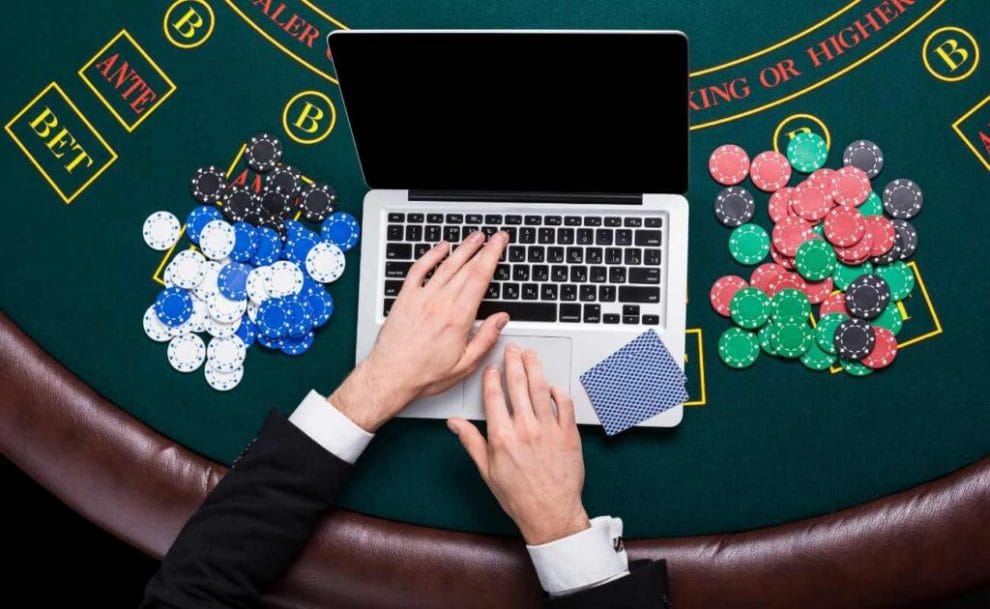 a top view of a person's hands typing on a laptop that is on a blackjack table with poker chips scattered on either side of the laptop and a pair of playing cards face down on the keyboard