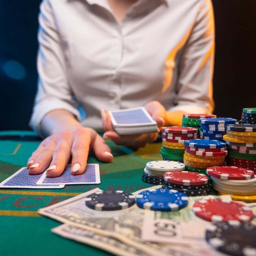 a woman dealer dealing two playing cards on a green blackjack table that has stacks of poker chips and money notes on it