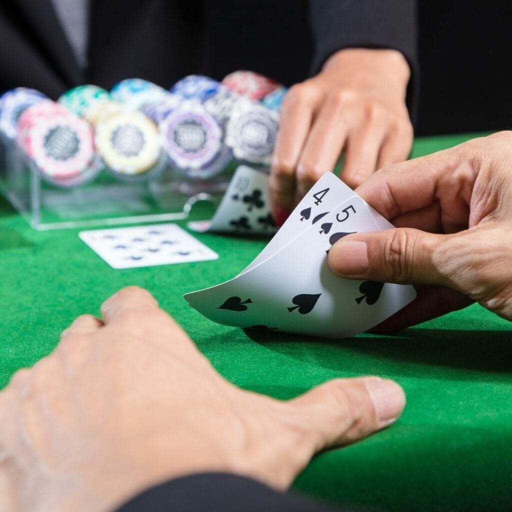 a person checking their hole cards, a four and five of spades, on a green felt poker table with the dealer and bank of poker chips in the background