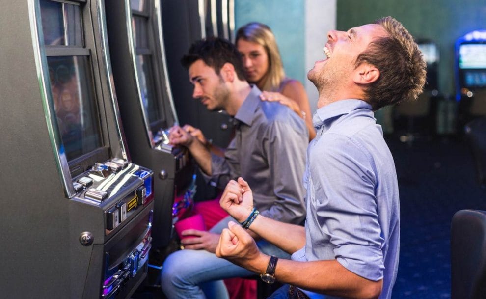 A man clenches his fists and throws his head back in victory before a slot machine.