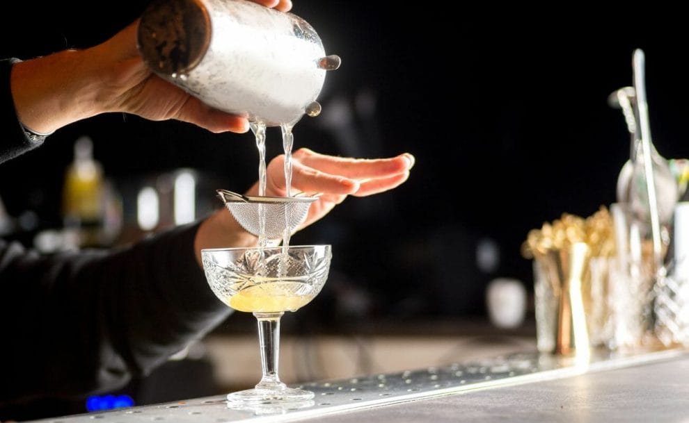 a bartender is pouring a cocktail into a crystal glass on the bar counter from a shaker and through a strainer