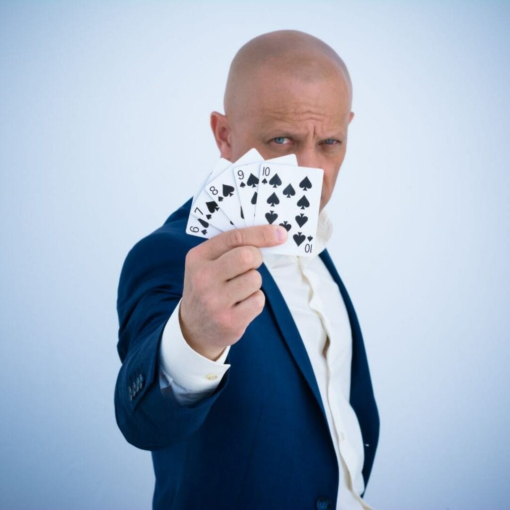 a man in a suit holding up a six to ten straight flush of spades in front of half his face