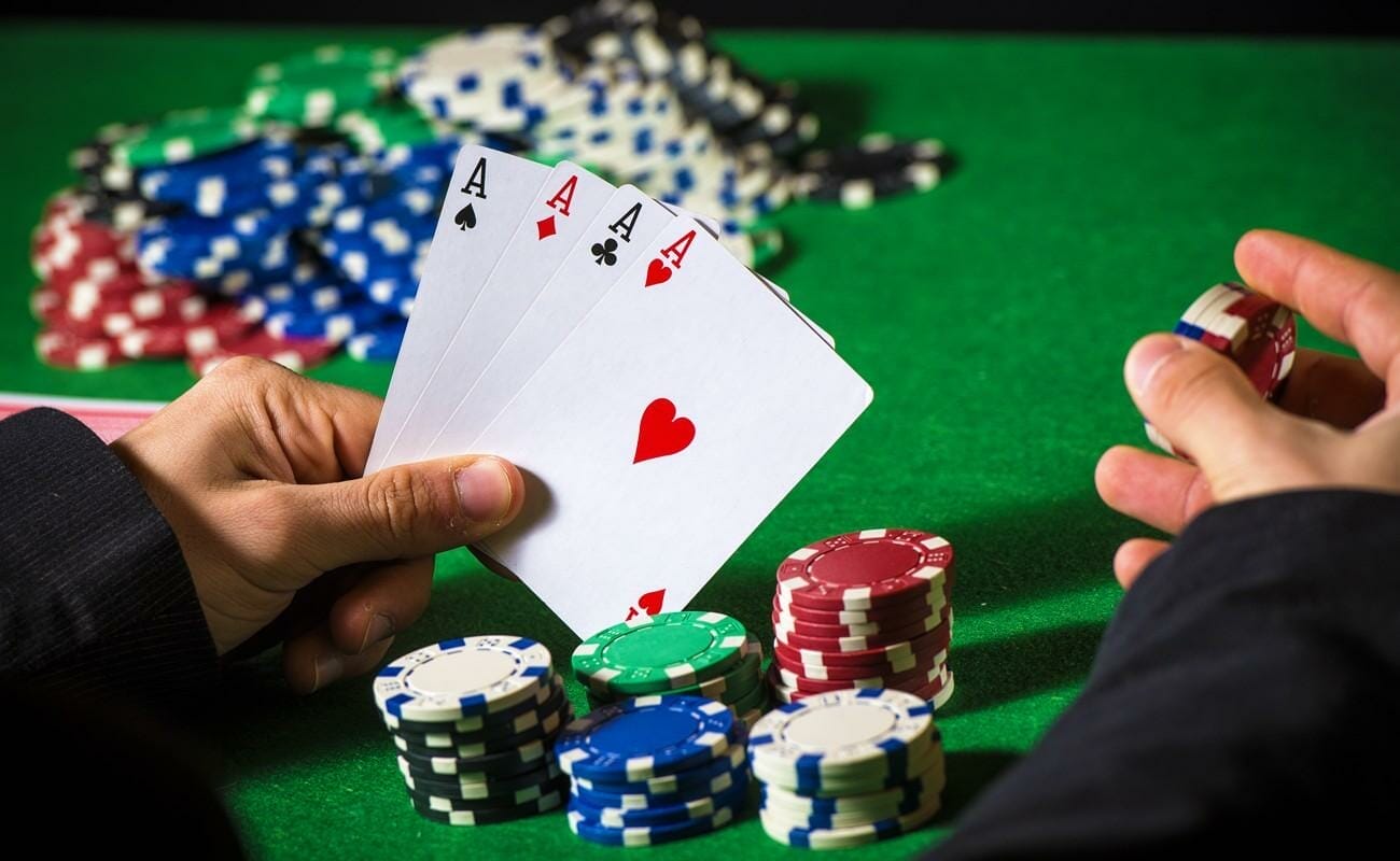 A person holds four aces in their left hand and a small stack of chips in their right. There is several stacks of chips between their two hands, and a large pot of chips in the background.