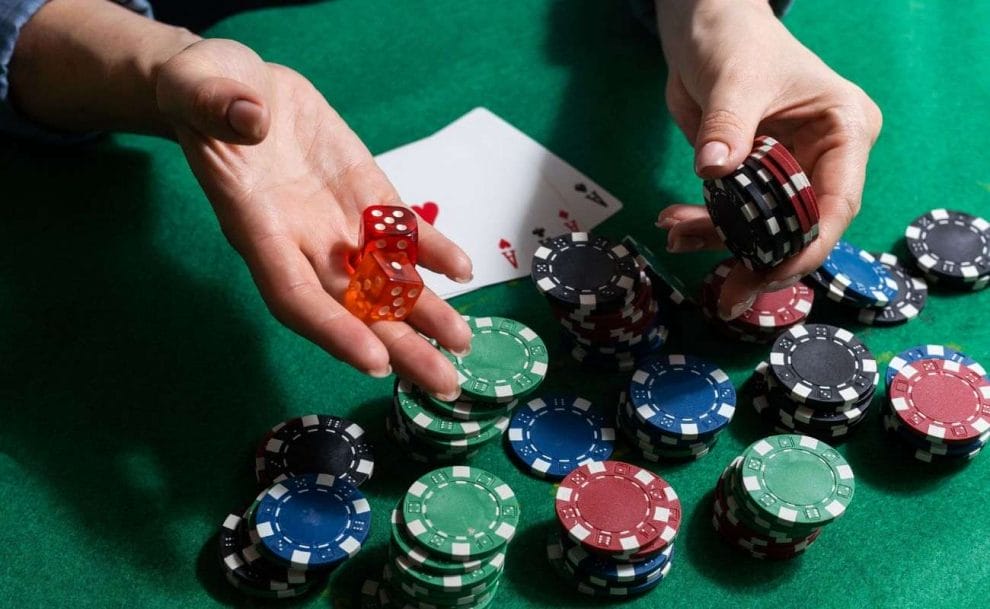 a person holding out a red pair of six-sided dice and poker chips over a green felt poker surface that has poker chips and four-of-a-kind ace playing cards on it 
