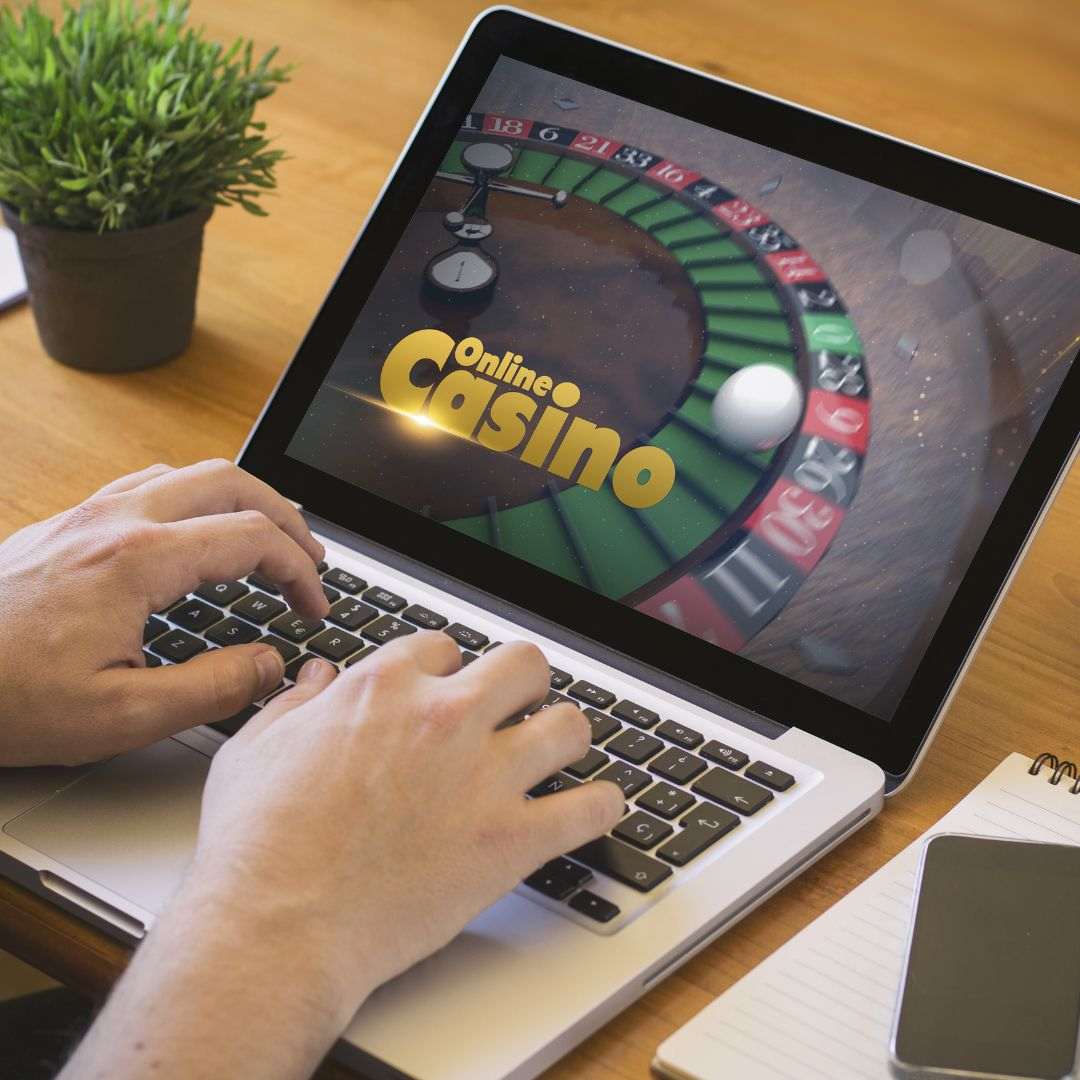 a close up of a person typing on a laptop displaying an online casino concept on the screen with a cellphone on a notepad beside the keyboard