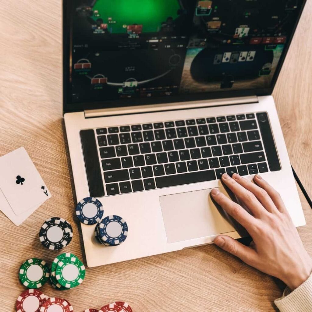 a person playing in an online multi-table poker tournament on a laptop on a wooden desk with playing cards and poker chips on it