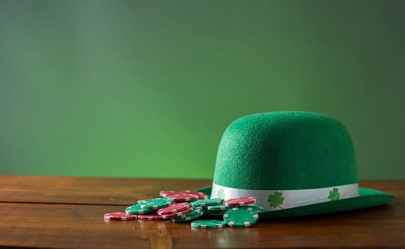 a green bowler hat with four leaved clovers on it on a wooden table next to a pile of red and green poker chips and a green background 