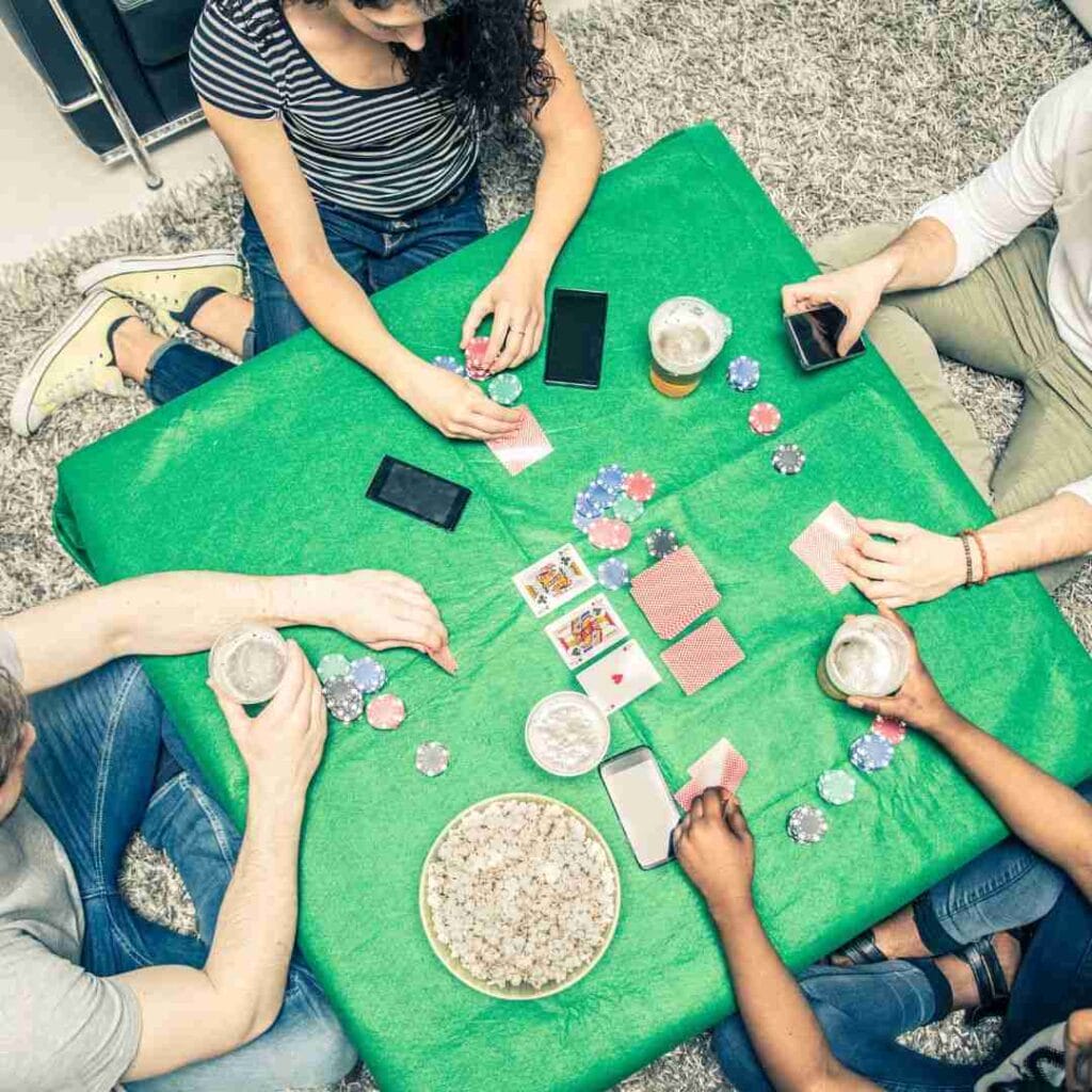 top view of four friends playing a game of poker at home on a DIY poker table that has playing cards, poker chips, cellphones, drinks and a bowl of popcorn on it