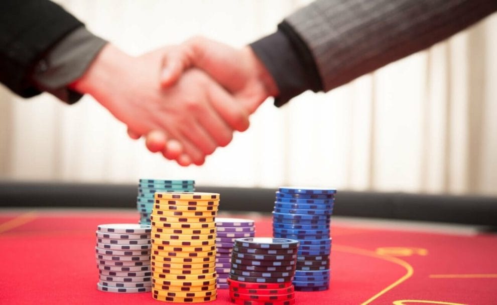 two people shaking hands above a red poker table that has multi-colored poker chips stacked on it 
