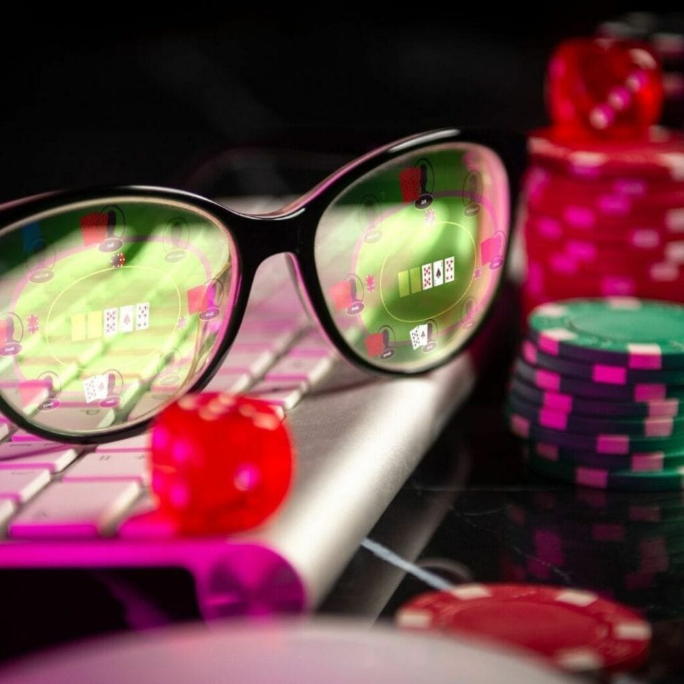 a pair of glasses on a computer keyboard reflecting a computer screen showing an online poker tournament in play, there are poker chips next to the keyboard and a red six-sided dice on the keyboard next to the glasses