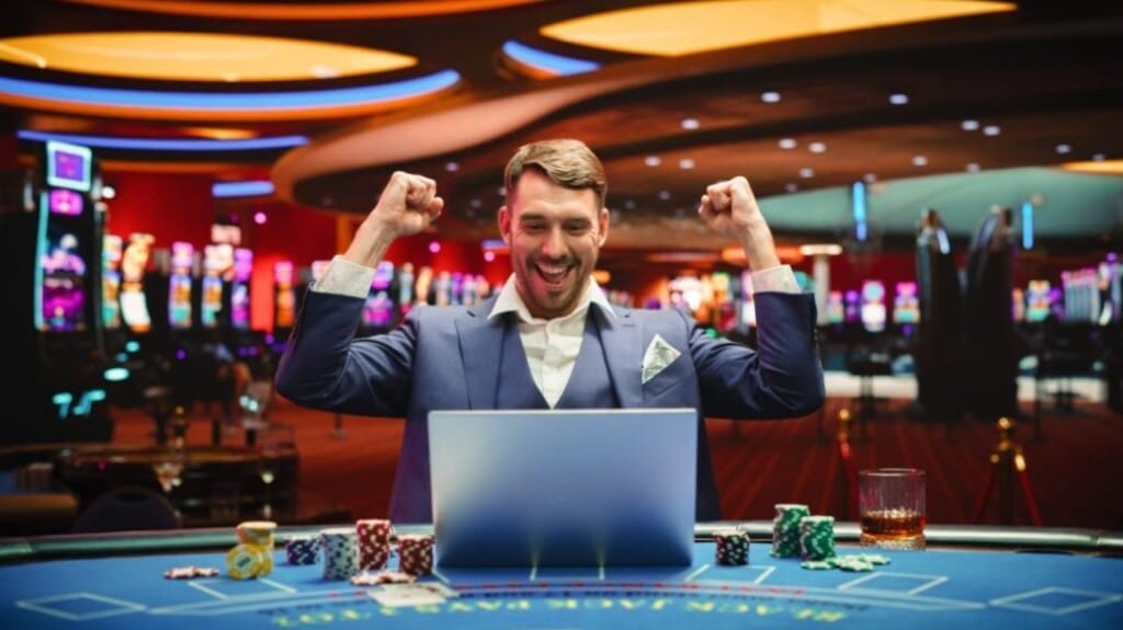 Man celebrating a big win while playing online casino games on a laptop, surrounded by poker chips and slot machines in the background.