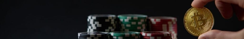 a person’s fingers holding a Bitcoin in front of stacks of poker chips