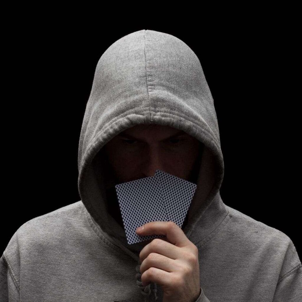 a man wearing a hoodie is holding up his two hole cards to cover his lower face during a game of poker