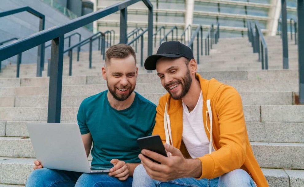 two men are smiling while sitting outside on some steps with a laptop and a cellphone