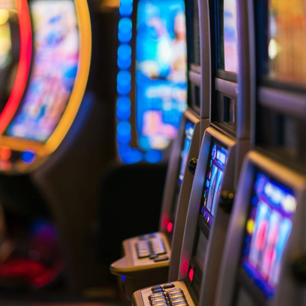 A side-view closeup of three slot machines lined up on a casino floor with the out-of-focus lights from other casino games in the background.