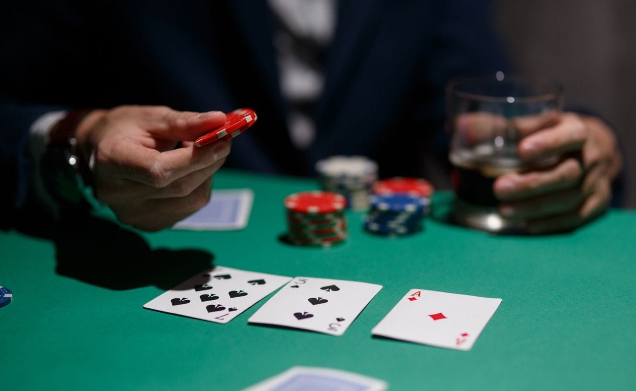 Close-up view of a person’s hands holding red poker chips above a green felt poker table. The table has playing cards and poker chips on it.