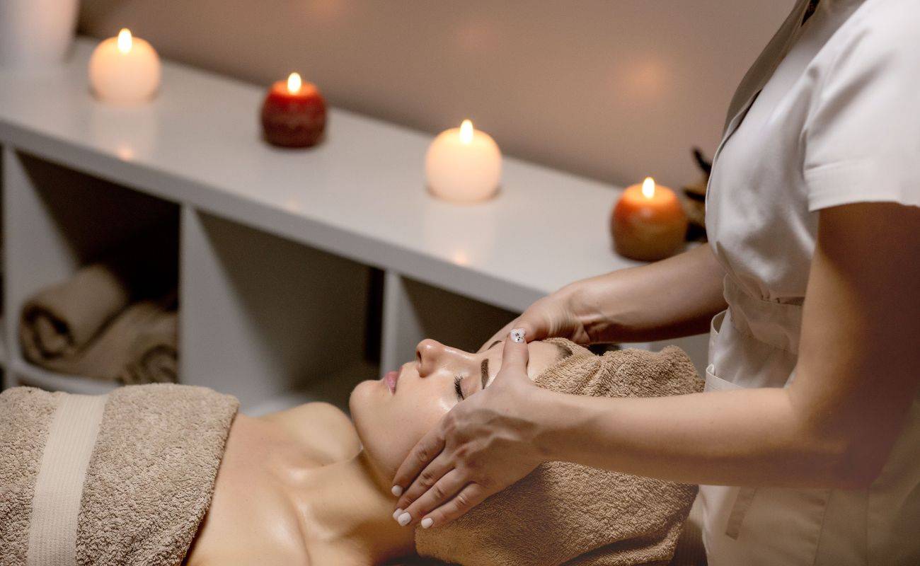 a woman is relaxing with a towel on her head and body while receiving a facial in a spa that has candles lit in the background