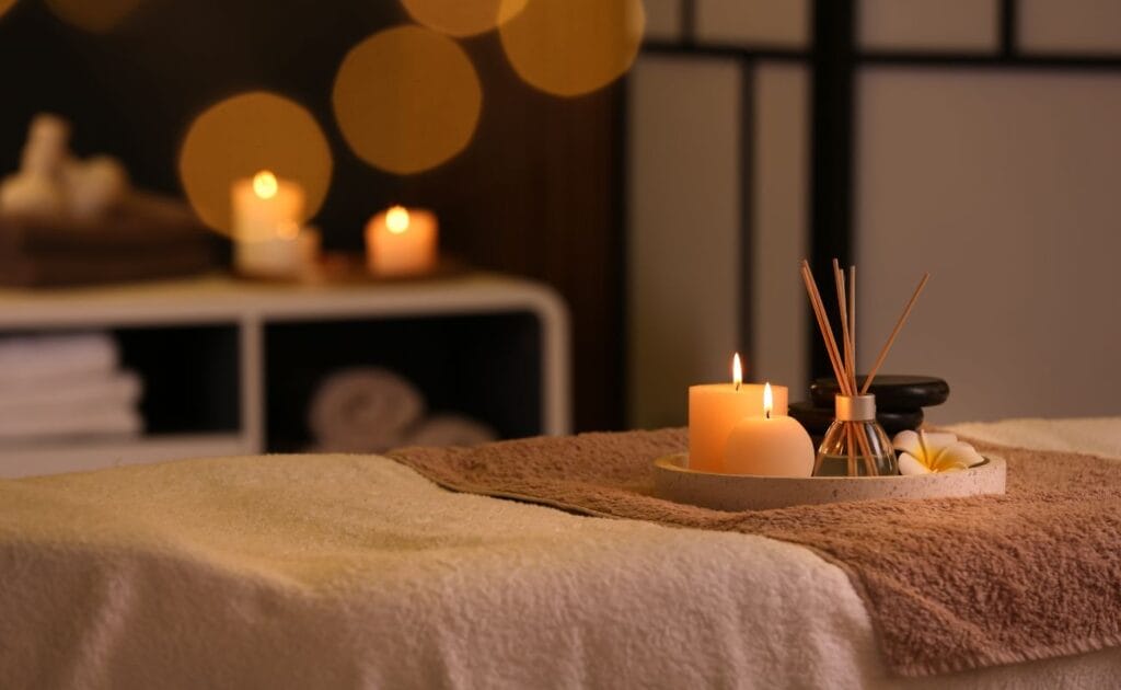 candles, a diffuser and hot stones on a tray on a spa bed in a spa with candles lit in the background