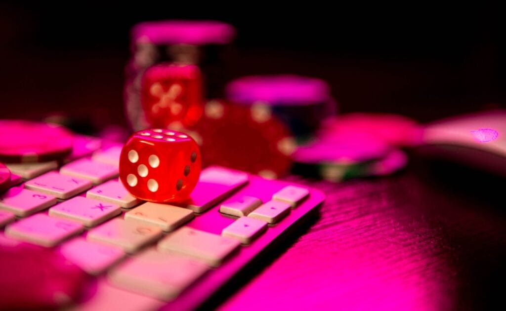 a red six-sided dice on a computer keyboard with poker chips blurred in the background in pink ambient lighting