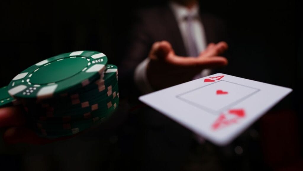 A close up of a man wearing a suit throwing an Ace of Hearts toward the camera, with a small stack of green poker chips to the left of the image.