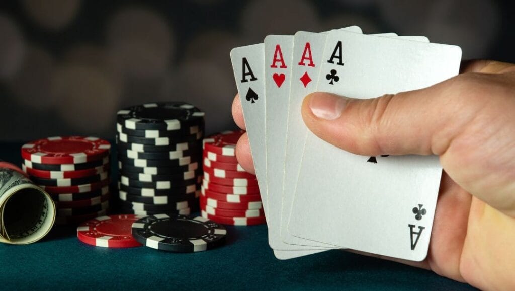 A close up of a poker player’s hand holding a four of a kind combination of Ace cards, with poker chips stacked in the background.