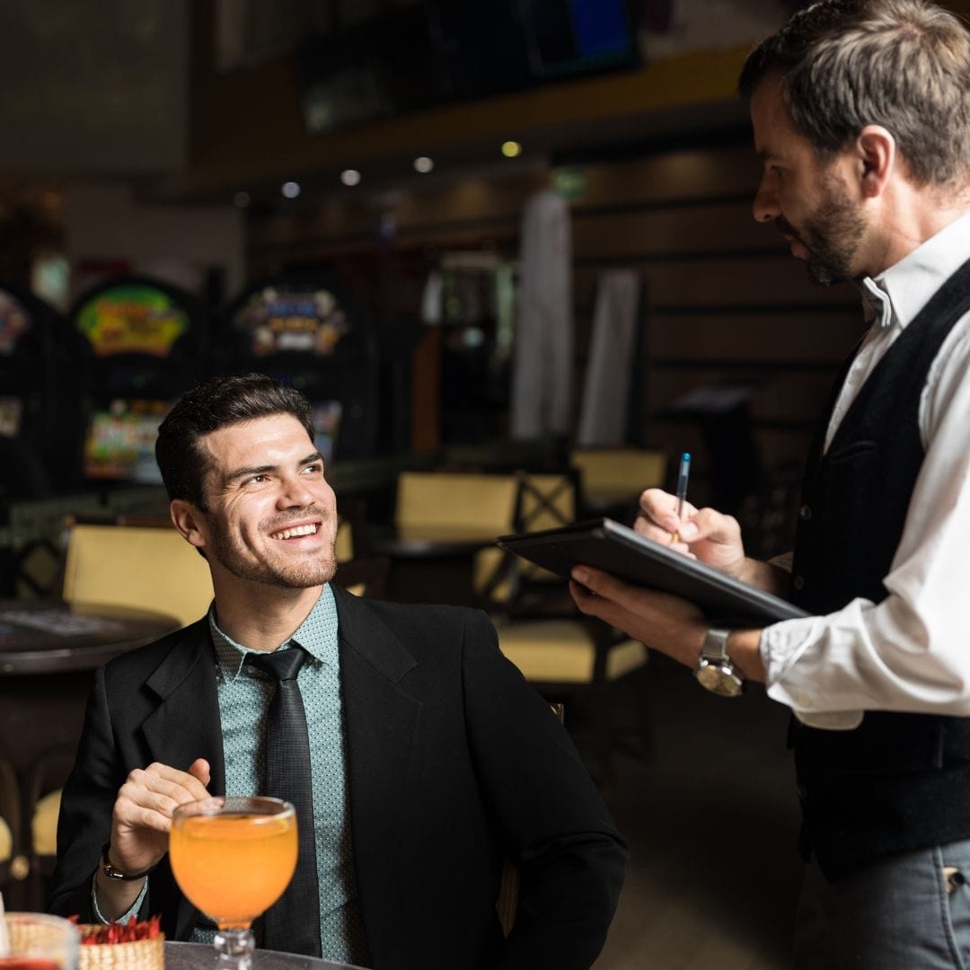 A waiter taking the order of a very happy man sitting at one of the tables at a casino restaurant.
