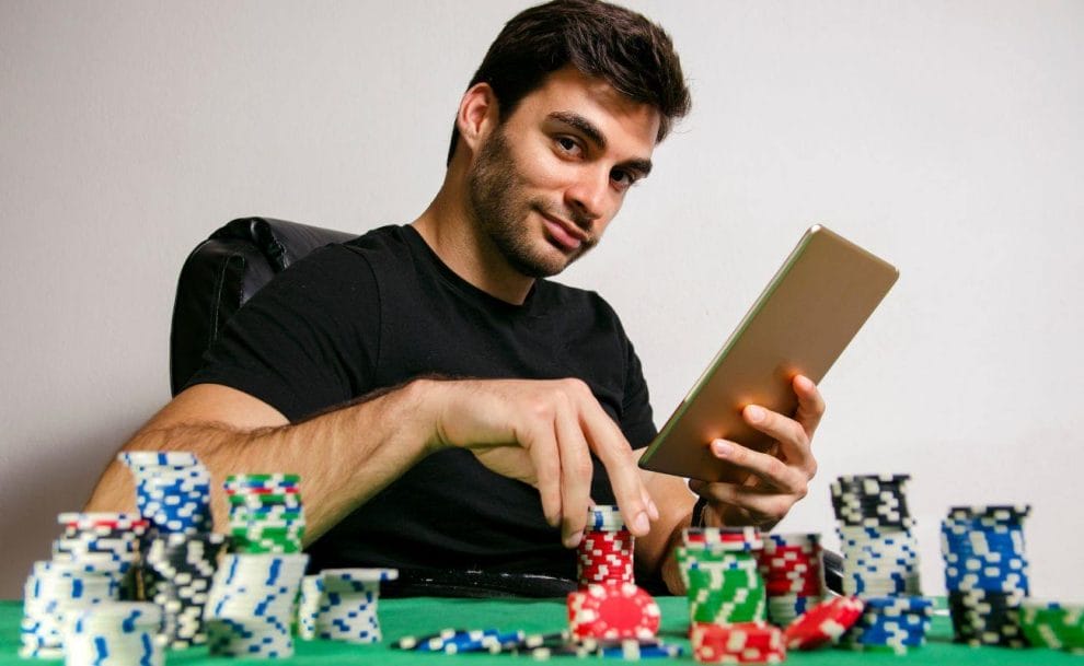 A man seated at a poker table, holding a tablet, with poker chips arranged on the table in front of him.