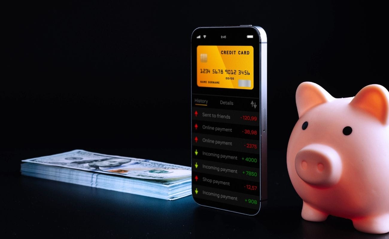 A stack of money and a pink piggy-bank on either side of a touch-screen cellphone showing incoming and outgoing online payments.