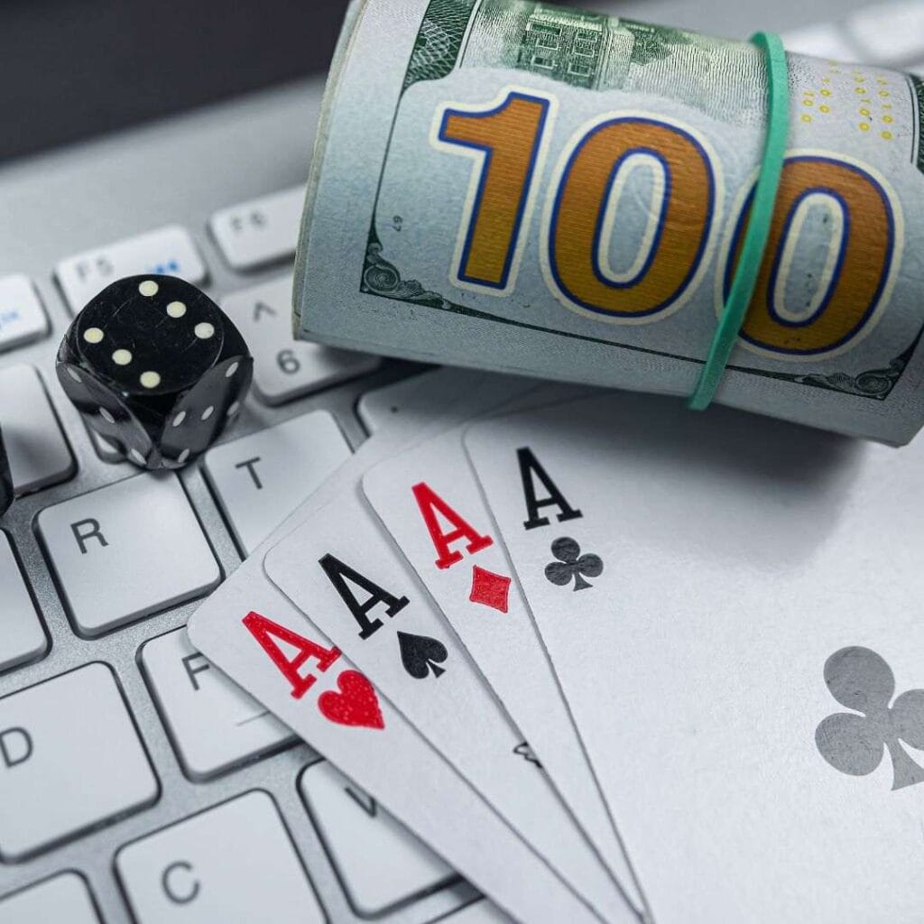 A rolled up stack of money, one black six-sided dice, and four of a kind Ace playing cards on top of a laptop keyboard.