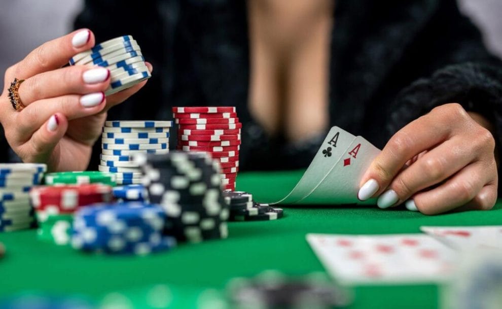 a woman shows her pocket pair of ace playing cards with one hand while picking up from a stack of poker chips with the other hand above a green felt poker table 