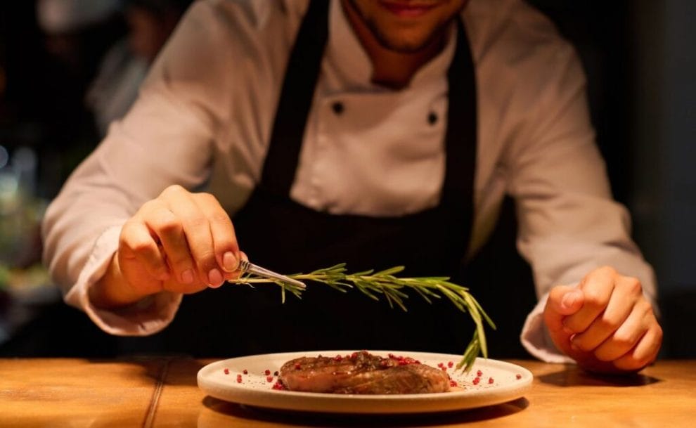 A chef garnishing a piece of steak on a white plate, with a stick of rosemary.